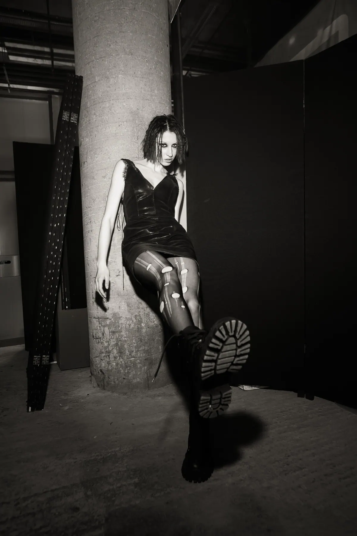 A model in a leather dress and heavy boots, one leg raised against a pillar