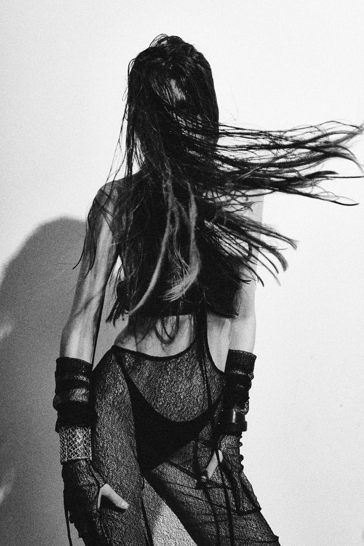 Black and white: a model seen from behind, hair thrown forward, mesh bodice and leather cuffs