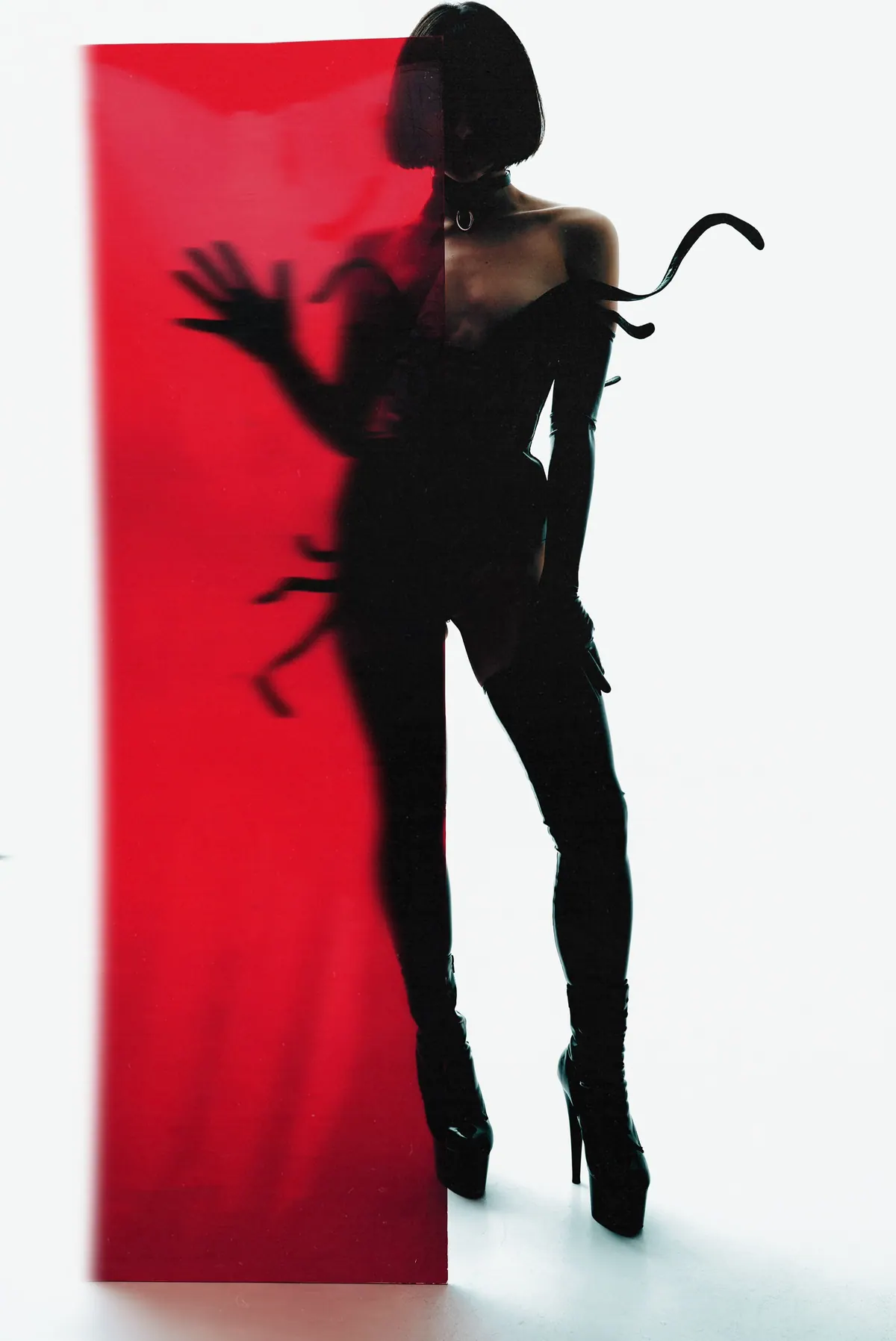 A figure in leather and thigh-high boots against a red panel, a shadow hand reaching from the left